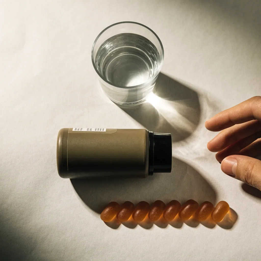 Un petit pot de complément sans inscription, à côté de quelques gommes et d'un verre d'eau sur fond papier clair